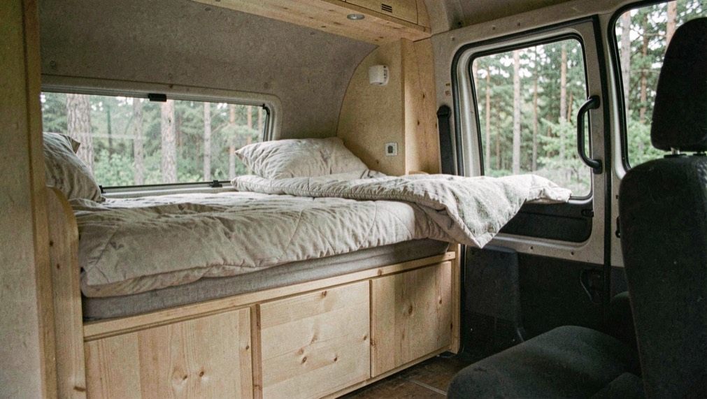 Interno di un furgone allestito con letto trasversale in legno chiaro, cassettone sotto, finestrino laterale aperto e vista su un bosco